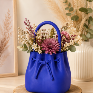 Sac décoratif violet avec anse rempli de fleurs variées, posé sur un rondin de bois.