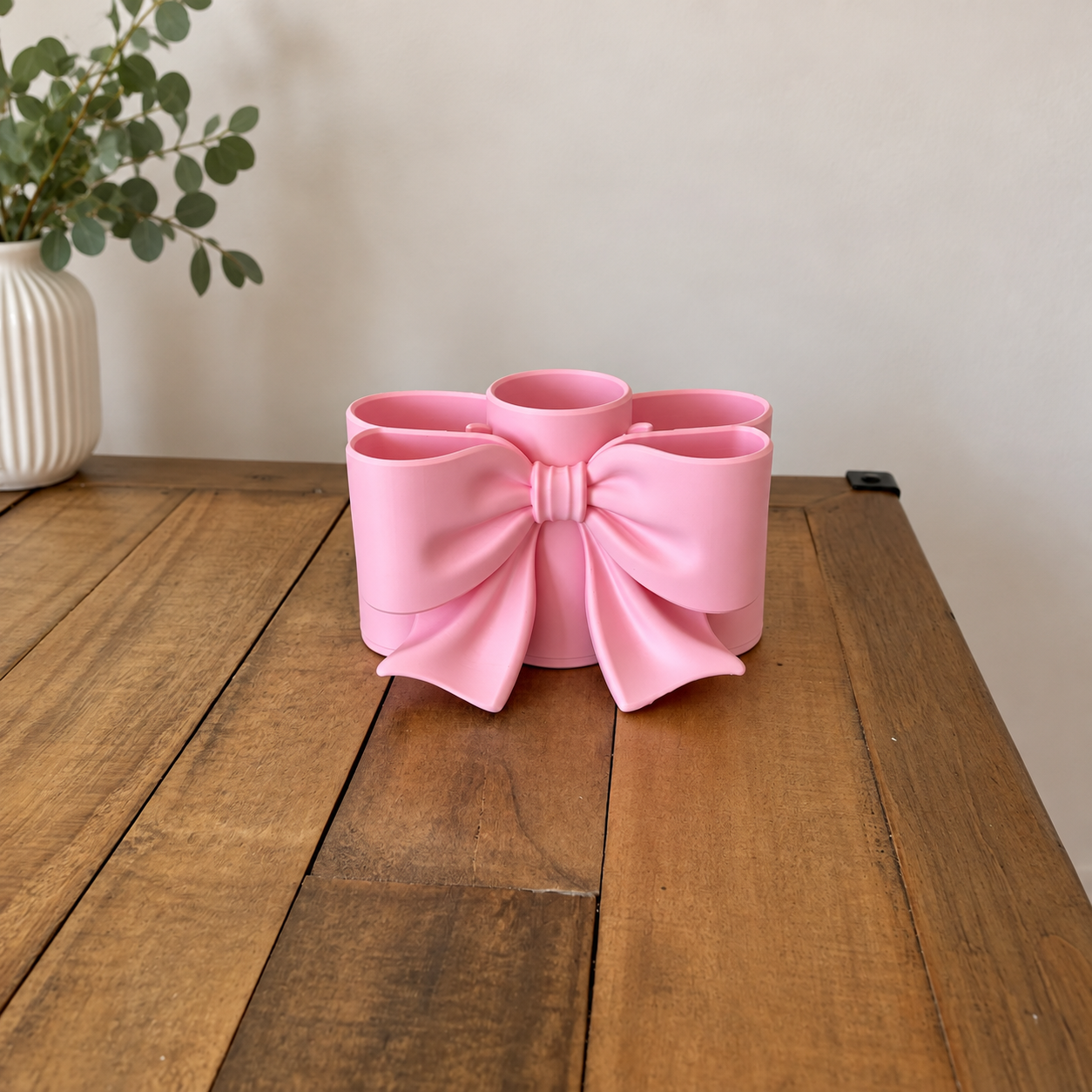 Porte-pinceaux de maquillage rose pastel en forme de nœud, posé sur une table en bois devant un mur beige